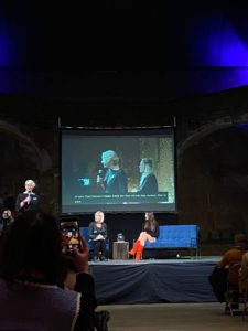 Professor Joanna Bourke standing on stage with a microphone, Jude Kelly sitting opposite Emily Ratajkowski. 